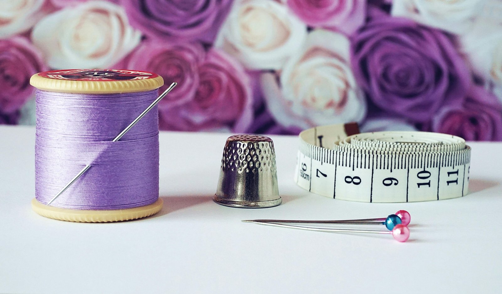 A close-up of sewing tools including lilac thread, a thimble, tape measure, and colorful pins with a rose backdrop.