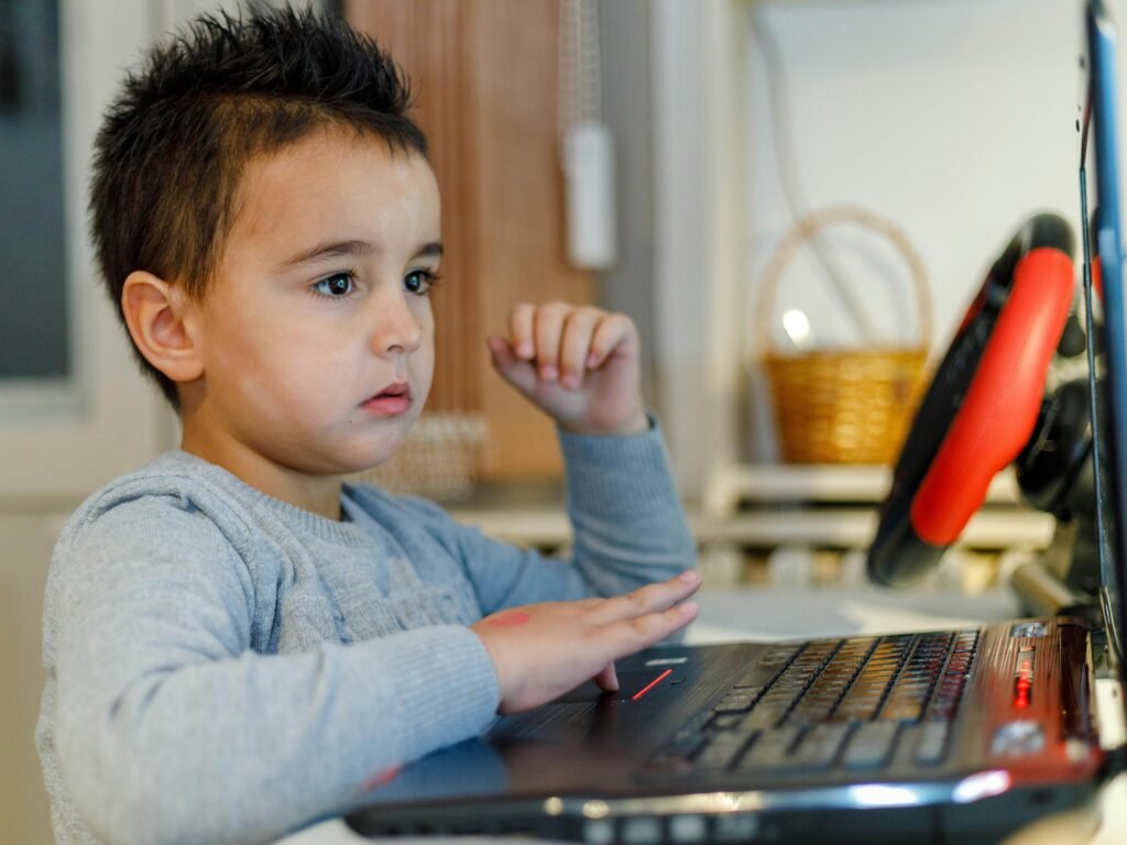 A young child focused on a laptop with a gaming steering wheel setup.